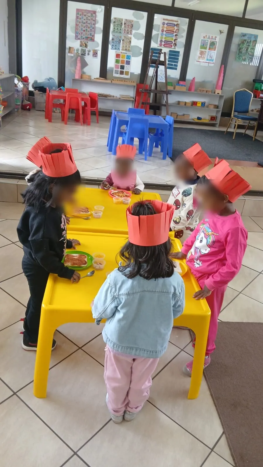 Little chefs in red paper crowns making pizza toast at two yellow tables in the classroom