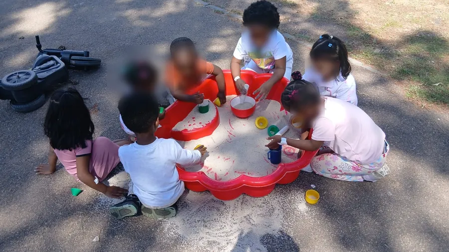 Young children around a red sandpit outdoors, scooping and pouring sand